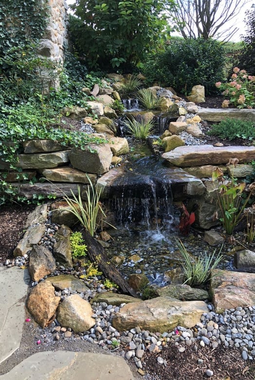 A garden waterfall cascading over mossy rocks framed by pink bleeding heart flowers and greenery