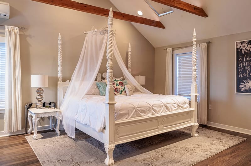 The cottage bedroom with a white four-poster canopy bed, exposed wood beams, and a sunlit sitting nook
